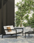 Modern patio with an Ethnicraft Jack Outdoor 2 Seater Sofa in black teak and off white, plus a brown accent pillow, sits by a low black coffee table. Green leafy plants grow against a white brick wall in the background.