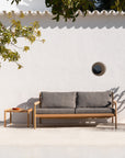 The Ethnicraft Jack Outdoor 2 Seater Sofa in FSC-certified teak with mocha cushions sits by a small wooden side table holding a glass of water and a dish, set against a white stucco wall with a round window and leafy shadows.