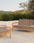 An outdoor patio features the Ethnicraft Jack Outdoor 2 Seater Sofa in FSC-certified teak with mocha Belgian fabric cushions, a beige blanket draped over the sofa, and green shrubs and hills in the background.