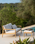 The Ethnicraft Jack Outdoor 2 Seater Sofa - Teak with Off White sits poolside, facing a footstool with a tray and glass of water. Lush greenery and trees fill the background beneath a clear sky.