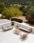 A modern patio showcases the Jack Outdoor 2 Seater Sofa in teak with off white by Ethnicraft, paired with matching teak furniture on a white floor. Surrounded by greenery, the space overlooks scenic hills.
