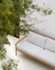 The Ethnicraft Jack Outdoor 2 Seater Sofa in teak with off white cushions sits on a cream patio, partially shaded by leafy green branches in the foreground.