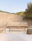 A modern patio setup with Ethnicrafts Jack Outdoor 3 Seater Sofa in teak and natural, matching armchair, teak coffee table, round ottoman, and a striped pillow on a light rug by a stone wall and tree, hills in the background.