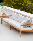 An Ethnicraft Jack Outdoor 3 Seater Sofa in teak with off-white cushions sits on a white patio beside a small side table topped with a woven vase, surrounded by greenery.