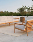 An outdoor patio features the Ethnicraft Jack Outdoor 3 Seater Sofa in teak with off-white cushions, two matching armchairs, a beige rug, large ceramic vase with greenery, sandals on the ground, and lush foliage under a clear sky.