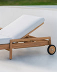 The Ethnicraft Jack Outdoor Adjustable Lounger - Teak features a reclining design, white cushioned seat and back, and two black wheels. Its shown on a smooth white surface with grass in the background.