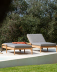 Two Ethnicraft Jack Outdoor Adjustable Loungers in teak with gray cushions sit poolside on a white patio. One features a rust pillow. Lush green grass and leafy trees enhance the tranquil setting.