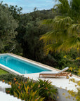 A rectangular swimming pool framed by greenery features the Ethnicraft Jack Outdoor Adjustable Lounger - Teak and palm trees nearby. The bright blue water stands out against the natural landscape on a sunny day.