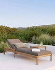 The Ethnicraft Jack Outdoor Adjustable Lounger - Teak, featuring brown cushions and two pillows, sits beside a small table with a drink and magazine on a patio bordered by green shrubs and hills beneath a clear sky.