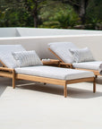 Two Ethnicraft Jack Outdoor Adjustable Loungers in teak, each with light gray cushions and patterned pillows, are arranged on a sunny patio with a small table and towel nearby. Trees in the background complete the inviting outdoor lounge.