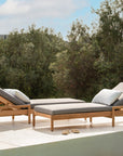 Two Jack Outdoor Adjustable Loungers in teak by Ethnicraft, with wheels and pillows, are set on a sunny patio by the pool. Sandals lie close, while lush green trees create a beautiful outdoor lounge setting.