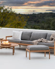A modern outdoor patio set with an Ethnicraft wooden sofa with gray cushions, a Jack Outdoor Footstool - Teak with Mocha by Ethnicraft, an outdoor lounge chair, two round tables, candles, plants, and a sunset view framed by trees.