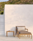 A minimalist outdoor patio features an Ethnicraft Jack Outdoor Footstool - Teak with Mocha, paired with a matching lounge chair and side table holding two glasses and a pitcher. A white staircase and wall contrast against green hills beyond.