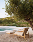 The Ethnicraft Jack Outdoor Footstool in Grade A Teak with off white cushions is shown on gravel under a leafy tree beside a pool, framed by lush greenery and hills. Part of the Ethnicraft Outdoor Collection.