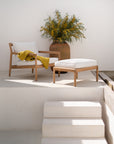 An Ethnicraft Jack Outdoor Footstool in teak with off-white cushions sits by a matching lounge chair on a white patio, complemented by a large brown vase of green foliage and yellow flowers, capturing the Ethnicraft Outdoor Collection’s elegance.