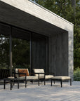 The Jack Outdoor Lounge Chair by Ethnicraft in Black Teak with Natural outdoor-grade cushions is paired with an ottoman and side table in neutral tones, set against a concrete building and surrounded by trees.