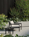 A modern outdoor lounge with an Ethnicraft Jack Outdoor Lounge Chair in Black Teak with Off White, paired with a small round table holding two glasses of water, surrounded by lush greenery and a dark wooden fence.