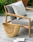 The Ethnicraft Jack Outdoor Lounge Chair in teak with mocha cushions sits on a patio, paired with a woven straw bag, an open book, and a closed notebook. Lush green plants complete this inviting outdoor scene.
