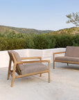 The Ethnicraft Jack Outdoor Lounge Chair - Teak with Mocha offers stylish comfort for your patio, set against scenic greenery and blue skies—perfect outdoor furniture for relaxing in style.