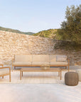 Serene patio with an Ethnicraft Jack Outdoor Lounge Chair in teak with natural upholstery, a beige sofa, coffee table, and two round ottomans on a neutral rug. Stone wall, tree, and hills complete the scene under a clear sky.