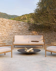 A modern outdoor lounge with two Ethnicraft Jack Outdoor Lounge Chairs in teak with off-white cushions and a wooden loveseat, set around a round fire pit by a stone wall, framed by a tree and rolling hills.