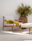 The Ethnicraft Jack Outdoor Lounge Chair in teak with off white cushions, draped with a yellow textured blanket, sits beside a matching ottoman. In the background is a large terracotta vase with leafy green branches and yellow flowers.