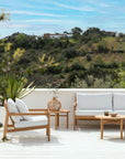 Outdoor patio with Ethnicraft Jack Outdoor Lounge Chairs in teak and off-white, plus a sofa and round coffee table. An inviting outdoor lounge set against green hills and blue sky.