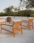 Two Ethnicraft Jack Outdoor Lounge Chairs in teak with off-white cushions sit on a patio beside a large ceramic planter and lush greenery, bathed in soft sunlight that enhances the serene, inviting vibe of this outdoor lounge space.