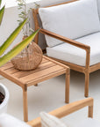 The Jack Outdoor Side Table - Teak by Ethnicraft holds a wicker vase with leafy branches, next to a matching teak sofa with light gray cushions. A green plant in a white pot appears partly visible in the foreground.
