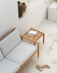A minimalist outdoor scene features a light wood Ethnicraft sofa with soft gray cushions beside the Ethnicraft Jack Outdoor Side Table in teak, which holds a glass of water and two books. Sandals lie nearby, and clay pots sit on a background ledge.