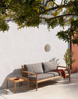 An Ethnicraft wooden outdoor sofa with gray cushions pairs with the Jack Outdoor Side Table - Teak. Against a white wall, a carafe, glass, brown throw, and cactus create a serene patio scene with tree-branch shadows.