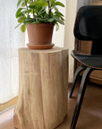 A potted plant rests on the SOBU Jupiter Side Table - Cedar, used as a side table beside a black chair and near a light curtain.