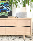 A SOBU Jupiter Sideboard - White Ash 4-door with wavy drawer fronts stands on a patterned rug. On top are a round wooden bowl and a potted plant in a gray and white pot, with an abstract painting hanging on the wall behind.