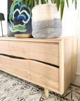 The SOBU Jupiter Sideboard - White Ash 4-door, featuring wavy handles, stands on a patterned rug. On top are a large gray-and-white planter with a leafy plant and a wooden bowl; a modern abstract painting is in the background.