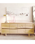 A SOBU Jupiter Sideboard in white ash with four doors stands on a rug, topped with two pink glass vases, a gold lamp, and white textured wall art. A large mirror leans against the wall to the right.