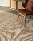 A wooden chair with a brown leather seat sits beside the Armadillo&Co Kalahari Rug in Natural / Chalk, handwoven from jute, in a minimalist room with a woven wooden cabinet and concrete floor.