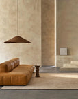 A minimalist living room with a tan leather sofa, Armadillo&Co’s Kalahari Rug in Natural/Chalk, a small brown side table, a large pendant light, and textured beige walls with a framed picture leaning on a step in the background.