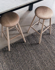 Two light wooden stools sit by a white marble table with black legs, atop Armadillo&Co’s Kalahari Weave Rug in Pewter/Charcoal, handwoven under Fair Trade practices and spread across the light wood floor.