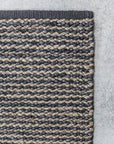 Close-up of the corner of the Armadillo&Co Kalahari Weave Rug in Pewter/Charcoal, highlighting its handwoven striped texture in gray and brown tones, laid on a light gray concrete floor.