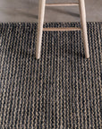 A light wooden chair sits on the Kalahari Weave Rug in Pewter/Charcoal by Armadillo&Co, handwoven from Fair Trade jute in alternating dark and light fibers, atop a light wood floor.