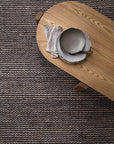 A wooden oval table with two grey ceramic plates and folded beige napkins sits atop the Armadillo&Co Kalahari Weave Rug in Pewter/Charcoal, a handwoven jute rug featuring dark and light stripes.