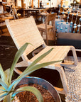 The Las Palmas Rocker by Noir Trading Inc.—a modern handcrafted woven lounge chair with a teak frame—is displayed beside a potted plant in a stylish furniture store with wooden tables, chairs, and decorative pieces in the background.