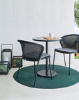 A modern outdoor seating area features two black Cane-line Lean Chairs and a round table on a green rug. A mug, tray, lanterns, and a plant accent the space against a light wall.