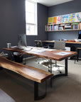 A modern office with a large live edge wooden desk, a SOBU Leviathan Bench in smoked acacia, gray walls, a bright window, shelves of books and colorful art, and two office chairs on carpeted flooring.