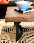 The Leviathan Live Edge Coffee Table by Moes, featuring solid acacia and a black metal base, displays a white bowl and two books. It rests on a textured black-and-white rug with a sofa and orange blanket behind it.