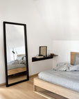 Minimalist bedroom with Ethnicraft’s Light Frame Floor Mirror in black, a light wooden bed topped with gray bedding, and a floating shelf for books, art, and decor. Crisp white walls and ceiling enhance the serene atmosphere.