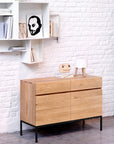 A modern room features the Ethnicraft Ligna Sideboard - 2 door with black metal legs, a white lamp, and an owl decor. White wall shelves above hold books and a black-and-white portrait against a white brick wall.