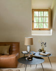 A cozy living room with a tan leather sofa, a small beige pillow, two round Luna Coffee Tables - Whiskey by Ethnicraft crafted by skilled artisans, a beige lamp, and a window with wooden trim letting in natural light.