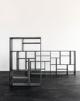 Two Ethnicraft M Rack Tall - Black shelving units stand on a dark wooden floor against a white wall, featuring open, geometric compartments in various sizes for a modern, minimalist look.