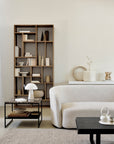 A modern living room with a curved beige sofa, black coffee table, marble side table with white lamp, minimal decor in neutral tones, and an Ethnicraft M Rack Tall - Teak bookshelf styled with books and vases.